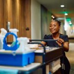 A housekeeping professional walks down a hallway pushing a housekeeping supplies cart.