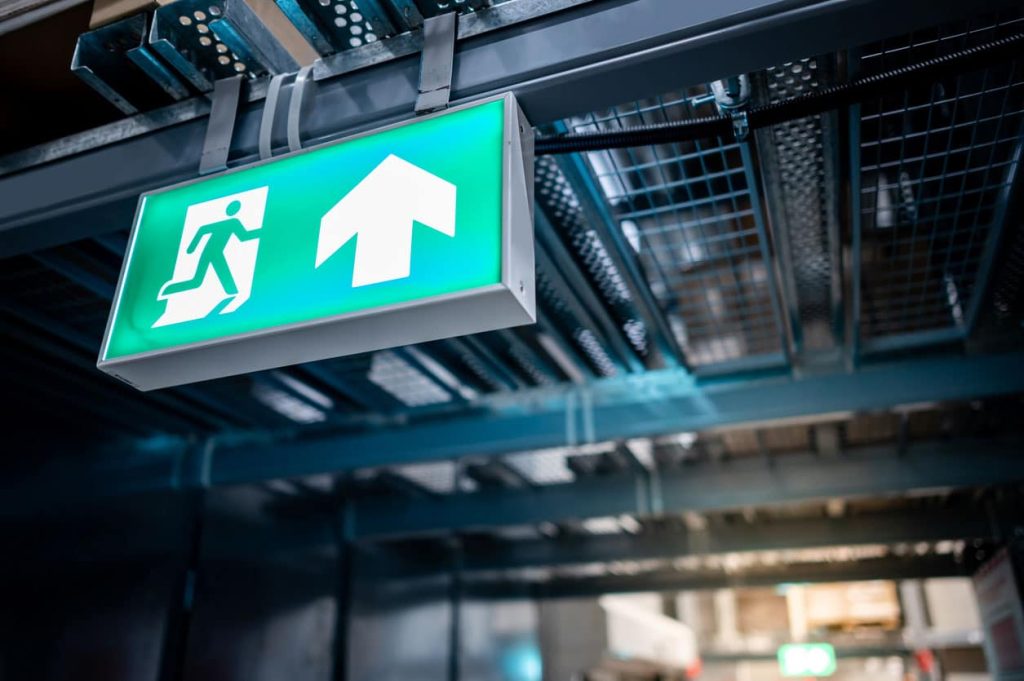 A fire exit sign is shown attached to overhead racking systems. The best warehouse safety tip is to ensure your fire exits are clearly signed and fully accessible. Learn more at 1stReporting.com.